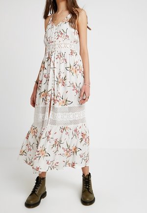 Mujer con vestido sin mangas blanco con flores y detalles de encaje, y botas verde oliva con cordones, de pie sobre un fondo blanco.
