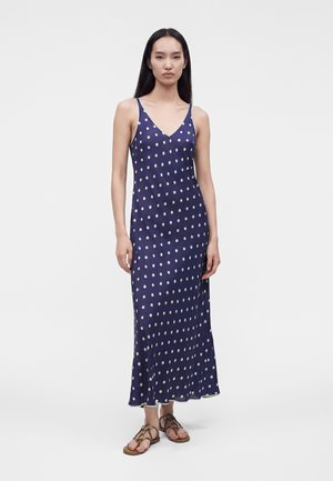 Femme debout portant une robe longue sans manches bleu marine à pois blancs et des sandales plates marron sur un fond uni.