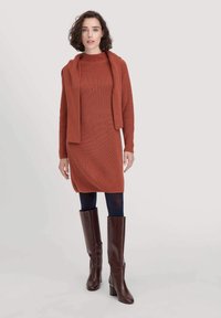Strickkleid in Rostfarbe mit einem Mock-Neck und kombiniert mit einer passenden langärmeligen Strickjacke. Das Model trägt kniehohe braune Stiefel mit Absatz.
