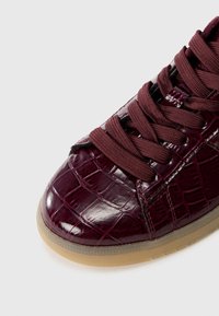 Baskets en cuir bordeaux avec un motif crocodile brillant, dotées de lacets épais couleur bordeaux et d'une semelle en caoutchouc beige. Vue rapprochée de la zone des orteils.