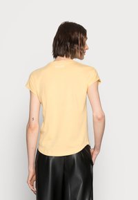 Persona con capelli corti castani, che indossa una maglietta gialla a maniche corte e pantaloni di pelle neri, in piedi con la schiena rivolta e la mano sinistra in tasca.