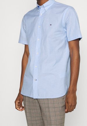 Mężczyzna w jasnoniebieskiej koszuli na krótki rękaw z małym logo na piersi oraz brązowych spodniach w kratę, stojący na jednolitym tle.