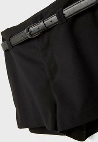 Shorts noirs sur mesure avec une ceinture fine en cuir noir passée dans les passants, sur un fond gris clair.