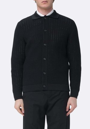 Uomo che indossa un cardigan nero texturizzato con bottoni sopra una camicia bianca col colletto e pantaloni neri, in piedi davanti a uno sfondo semplice.