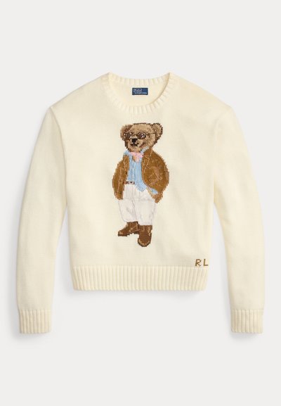 Ιβουάρ πουλόβερ με πλεκτή εικόνα αρκούδας που φοράει καφέ σακάκι και γυαλιά, με ανοιχτό γαλάζιο πουκάμισο και λευκό παντελόνι.