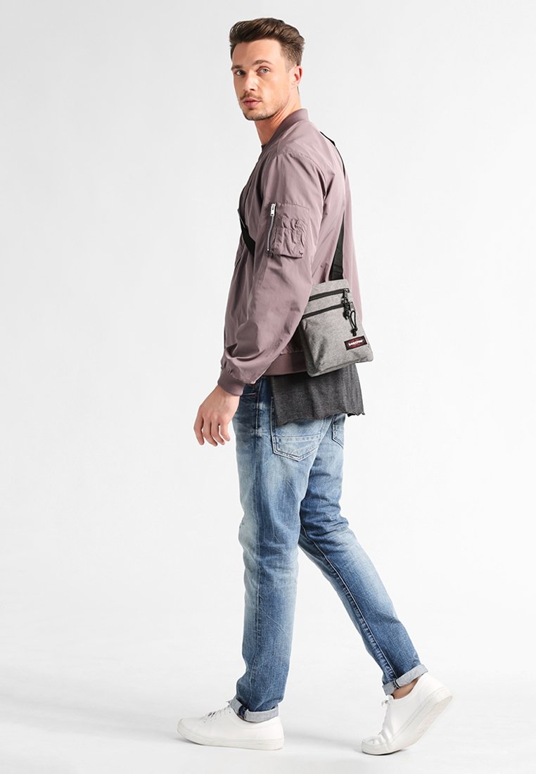 Borsa a tracolla grigia con due scomparti con zip, indossata sopra una giacca viola e sovrapposta a maglie nere e grigie, abbinata a jeans blu e sneakers bianche.
