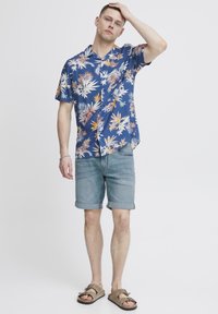 Chemise hawaïenne bleue à manches courtes avec un motif floral ; associée à un short en denim retroussé et des sandales marron. Le modèle se tient avec une main sur la tête.