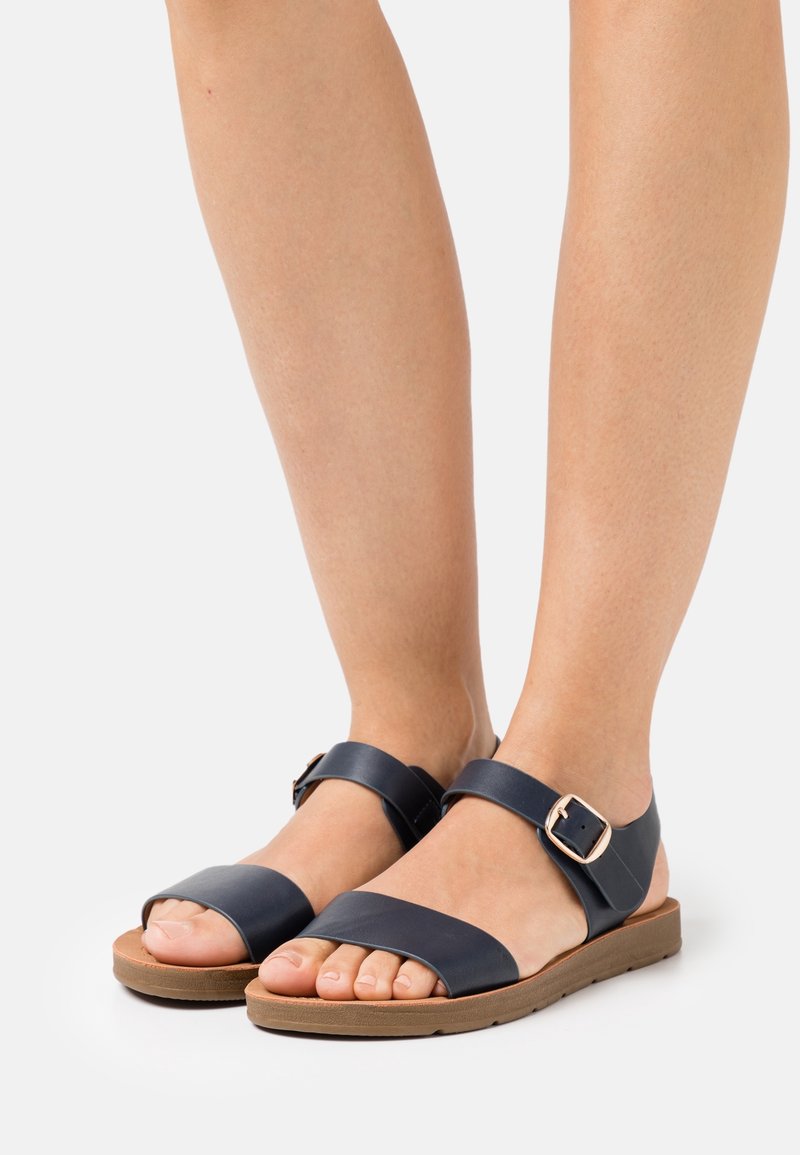 Pieds portant des sandales en cuir noir avec une bride à boucle, une semelle plate de couleur beige et les orteils découverts sur un fond gris clair.