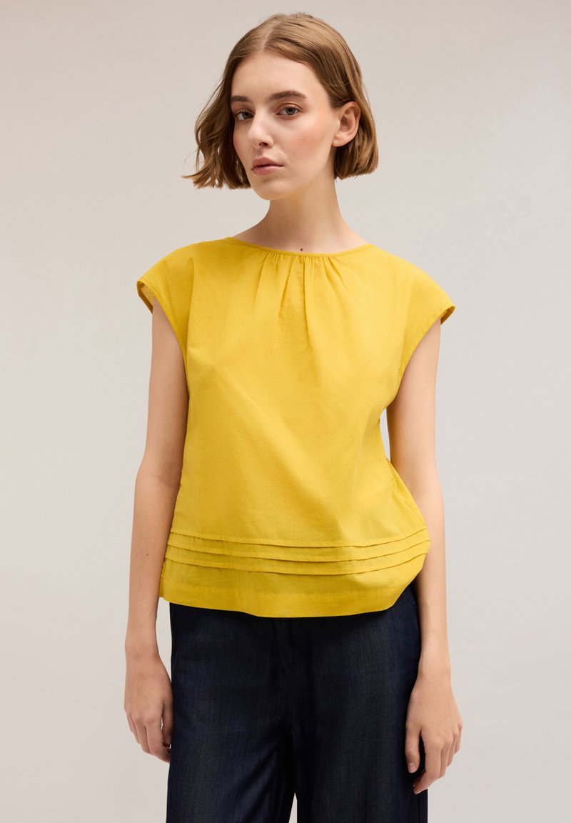 Top giallo a maniche corte con scollo arricciato e dettagli plissettati all'orlo, realizzato in tessuto leggero. Indossato con pantaloni in denim scuro.