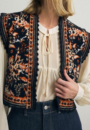 Vrouw draagt crèmekleurige blouse met striksluiting bij de hals, donker gebloemd gebreid vest en donkerblauwe high-waisted denim jeans, hand houdt zoom van het vest vast.