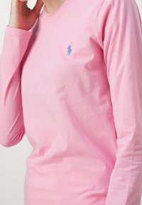 Långärmad rosa T-shirt i bomull med en blå broderad logotyp på bröstet. Mjuk textur med rund halsringning.