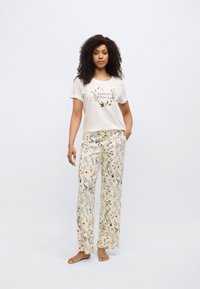 Μπεζ βαμβακερό t-shirt με φλοράλ τύπωμα και κείμενο. Τα ελαφριά παντελόνια πιτζάμας που ταιριάζουν έχουν ένα φλοράλ μοτίβο σε κίτρινο και πράσινο πάνω σε ανοιχτόχρωμο φόντο.