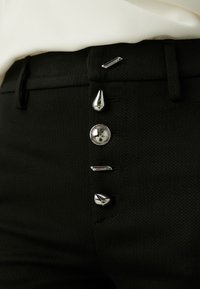 Schwarze strukturierte Hose mit fünf metallischen Knöpfen in verschiedenen Formen entlang der Vorderseite, darunter rund, tropfenförmig und stabförmig.