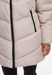 Ljus beige pufferjacka med dragkedja fram, diagonal quiltad mönstring, sidficka och svart dragkedjehårdvara. Mjuk, slät textur.