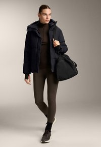 Čierna pufrová bunda s kapucňou, skombinovaná s hnedým topom a legínami. Obsahuje čiernu športovú tašku. Vyznačuje sa elegantným dizajnom a textúrovanou tkaninou.