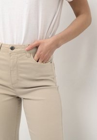 Pantaloni beige slim-fit con una texture liscia, caratterizzati da tasche frontali, chiusura a bottone e dettagli di cucitura visibili. Indossati con una maglietta bianca.