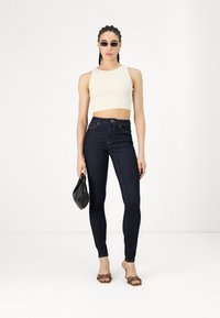 Top corto crema, jeans de mezclilla oscuros de cintura alta, bolso negro y sandalias de tacón marrón con correas decorativas. Estilo minimalista.
