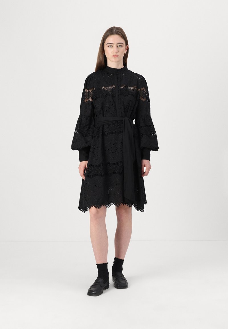 Vestito nero con maniche lunghe a sbuffo e colletto alto, caratterizzato da dettagli in pizzo, una cintura con nodo e un orlo scalloppato. Tessuto testurizzato.