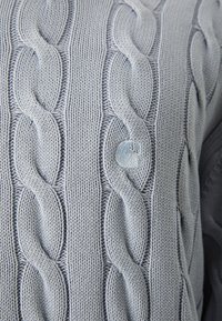 Hellblauer Strickpullover mit einem Kabelmuster und einer strukturierten Oberfläche sowie einem runden dekorativen Knopf auf der Vorderseite.