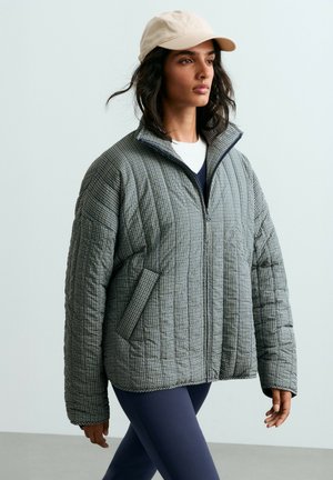 Femme portant une veste matelassée à carreaux, des leggings bleu marine, une chemise blanche et une casquette de baseball beige marchant sur un fond gris clair.
