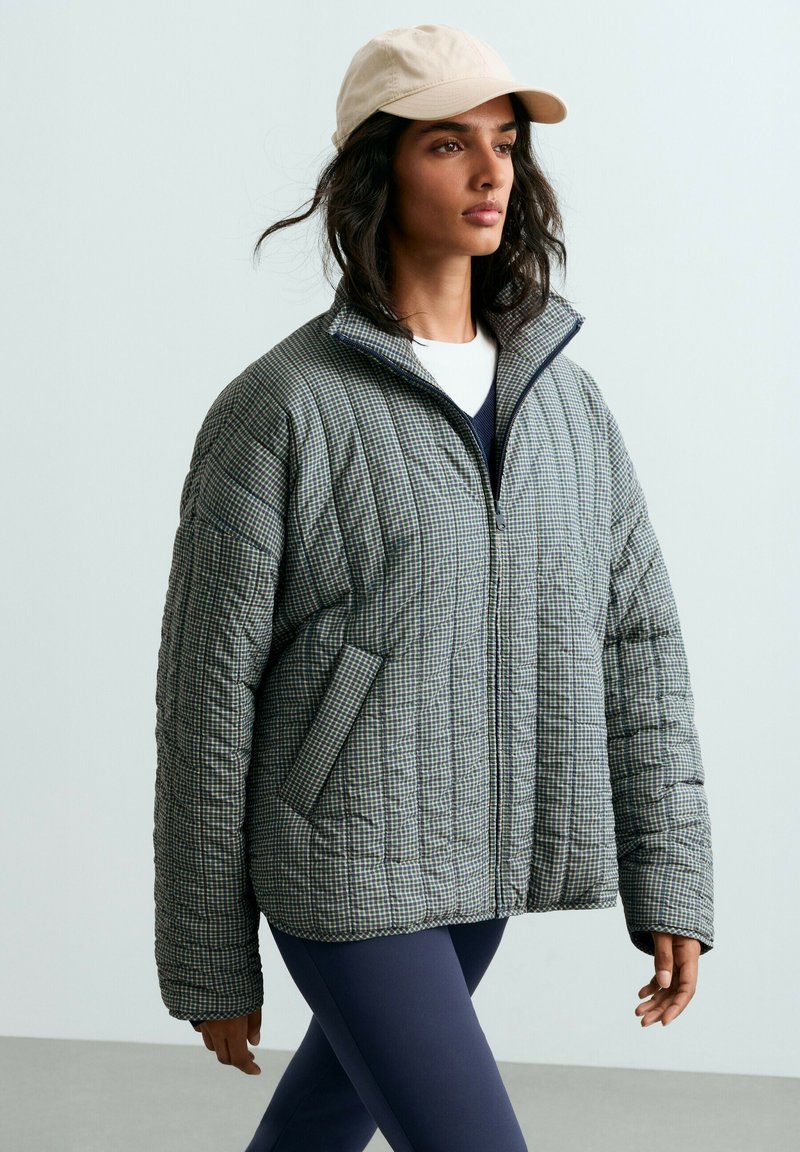 Femme portant une veste matelassée à carreaux, des leggings bleu marine, une chemise blanche et une casquette de baseball beige marchant sur un fond gris clair.