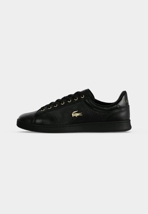 Baskets basses en cuir noir avec œillets dorés et logo crocodile Lacoste sur le côté, présentées sur un fond gris uni.
