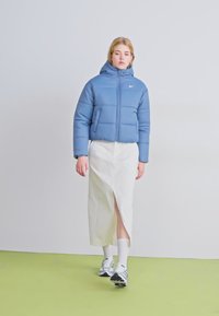 Chaqueta de plumas azul claro con capucha, cierre de cremallera y logo de Nike, combinada con una falda blanca con una abertura frontal y zapatillas grises.
