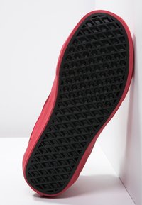 Suela roja de zapatilla con un fondo de goma negra texturizada que presenta un patrón geométrico, diseñado para ofrecer tracción y durabilidad.