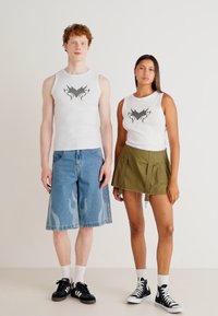 Du modeliai stovi šalia vienas kito. Vyrų modelis vilki baltą marškinėlį su juodu piešiniu ir mėlynus džinsinius šortus su ugnies raštais. Moterų modelis taip pat vilki baltą marškinėlį su panašiu piešiniu ir alyvuogių žalumo plisuotą sijoną. Abu modeliai dėvi baltas kojines, o vyrų modelis turi juodus sportinius batelius.