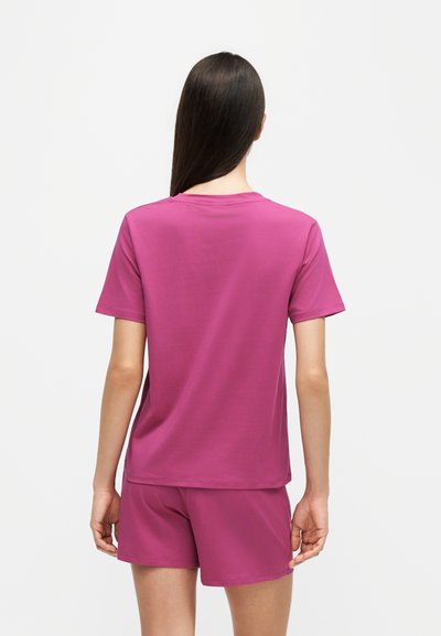 Femme aux longs cheveux foncés, debout de dos, portant un t-shirt uni à manches courtes assorti et un short de couleur magenta vif.