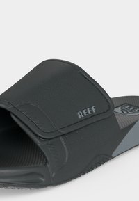 Sandale slide noire réglable avec semelle texturée et logo «REEF» sur la bride, conçue pour un usage décontracté.