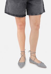 Bailarinas de cuero gris con punta afilada, cintas de tobillo entrelazadas y detalles plisados, combinadas con pantalones cortos de mezclilla negros.