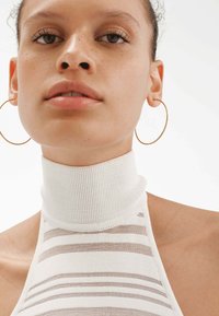 Top halter blanco con cuello alto, con rayas horizontales en color beige, textura acanalada y grandes pendientes de aro dorados.