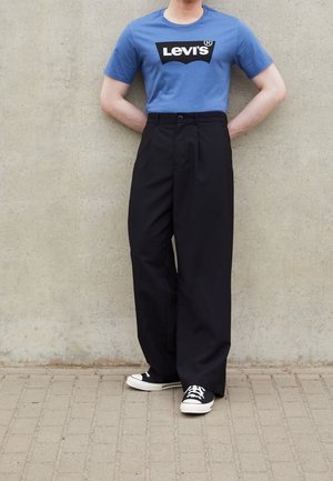 Personne portant un t-shirt Levi’s bleu, un pantalon large noir taille haute et des baskets noir et blanc, debout contre un mur en béton.