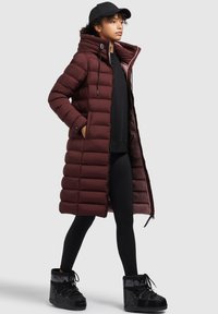 Doudoune bordeaux avec capuche et fermeture éclair, dotée d'un design matelassé. Portée sur des leggings noirs et associée à des bottes noires à lacets.