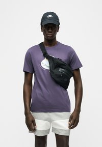 Άνδρας που φοράει μοβ μπλουζάκι με γραφικά, γκρι και λευκά σορτς, μαύρο καπέλο Nike και μαύρη τσάντα χιαστί μπροστά σε απλό λευκό φόντο.