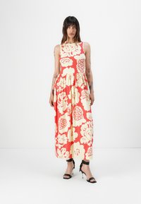 Maxi dress in coral with large yellow floral print. Sleeveless design with a fitted bodice and flowing skirt. Paired with black strappy heels.