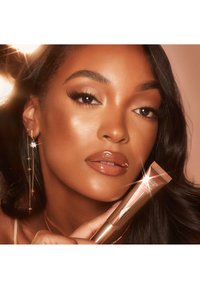 Kosmetikprodukt in einer roségoldenen Tube, die von einer Person gehalten wird. Zeigt glänzende Lippen, lange Wimpern und glitzernde silberne Ohrringe. Weicher, warmer Hintergrund.