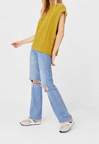 Haut jaune tricoté sans manches avec motif torsadé, associé à un jean bleu clair déchiré et à des baskets beiges avec semelles noires.