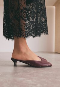 Scarpe mule in pelle bordeaux, con punta affusolata, piccoli perni dorati lungo i lati e tacco in legno basso.