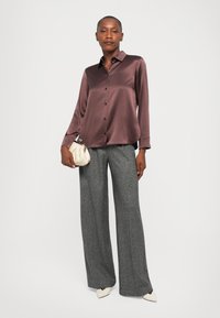 Donna che indossa una camicia lucida marrone con bottoni, pantaloni grigi a gamba larga, scarpe bianche con punta e tiene in mano una piccola borsa beige.