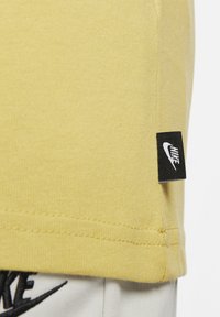 Tricou din bumbac galben deschis cu etichetă Nike neagră pe lateral, având o textură fină și mâneci scurte.