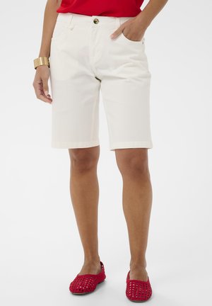 Osoba nosiaca biele šortky po kolená, červený top, červené tkané nazúvacie topánky a zlatý náramok na zápästie, stojaca s jednou rukou v vrecku.