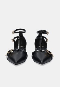 Tacones de cuero charol negro con punta afilada, tiras en el tobillo y acentos de hebilla dorada; diseño elegante con textura suave.