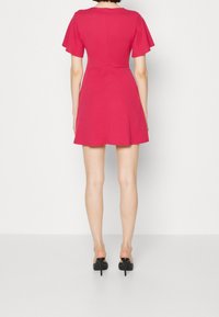 Dopasowana sukienka z krótkimi rękawami w jaskrawym różu, rozkloszowany dół, gładki materiał, okrągły dekolt i zamek błyskawiczny z tyłu. Noszona z czarnymi szpilkami.