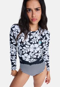 Γυναίκα που φοράει μακρυμάνικο μαύρο rash guard με λευκά λουλουδάτα σχέδια και μαύρο-άσπρο ριγέ μαγιό κάτω μέρος, στέκεται μπροστά σε ένα μονόχρωμο φόντο.