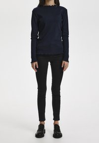 Granatowa bluzka z długim rękawem o prążkowanej strukturze i falbaniastych brzegach, w połączeniu z czarnymi dopasowanymi dżinsami i czarnymi loafersami.