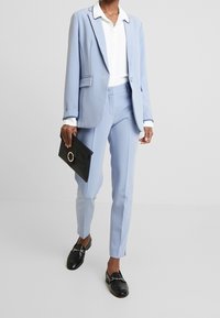 Donna che indossa un completo blu chiaro con camicia bianca, tiene una pochette nera e indossa mocassini neri, in piedi su uno sfondo neutro.