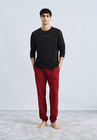 Cămașă neagră cu mâneci lungi cu textul "Calvin Klein"; pantaloni de pijama roșii cu un model mic de stele aurii; croială lejeră și manșete elastice.