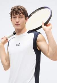 Camisola atlética branca sem mangas com detalhes em azul-marinho, apresentando o logótipo EA7. O modelo segura uma raquete de ténis preta numa pose neutra.